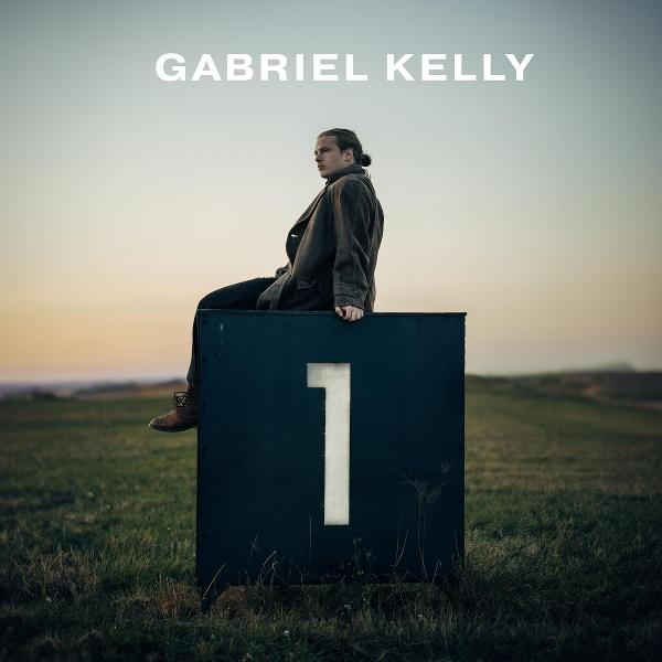 Ein Mann sitzt auf einer Kiste mit einer '1'. Im Hintergrund sind Feld und Himmel, darüber steht 'Gabriel Kelly'.