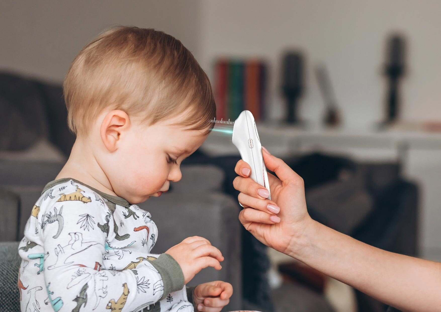 Dziecko ma mierzoną temperaturę białym termometrem. Ręka trzyma urządzenie blisko czoła. Dziecko ma na sobie koszulę z nadrukiem dinozaura.
