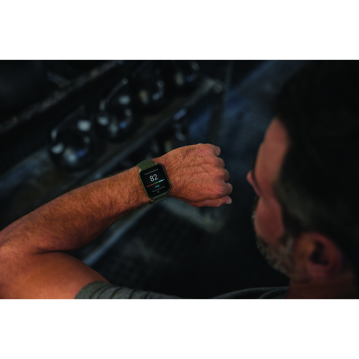 Man met smartwatch controleert trainingsbereidheid, 82. Achtergrond: gymtoestellen, gewichten. Focus op pols.