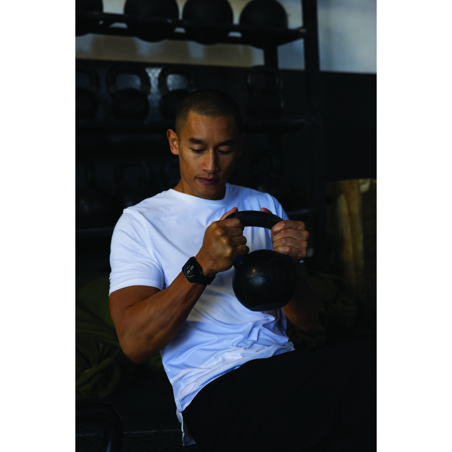 Man in wit shirt houdt zwarte kettlebell vast. Hij heeft een horloge. Achtergrond heeft kettlebells op een rek.