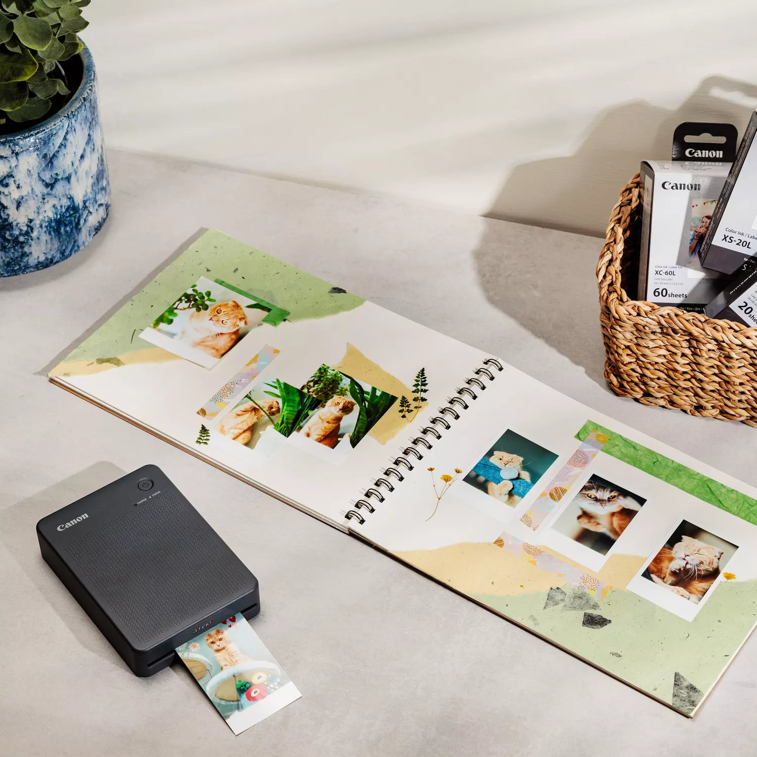 Drukarka Canon drukuje zdjęcie. Otwarty album z fotografiami kotów leży na stole, obok tuszu i rośliny.