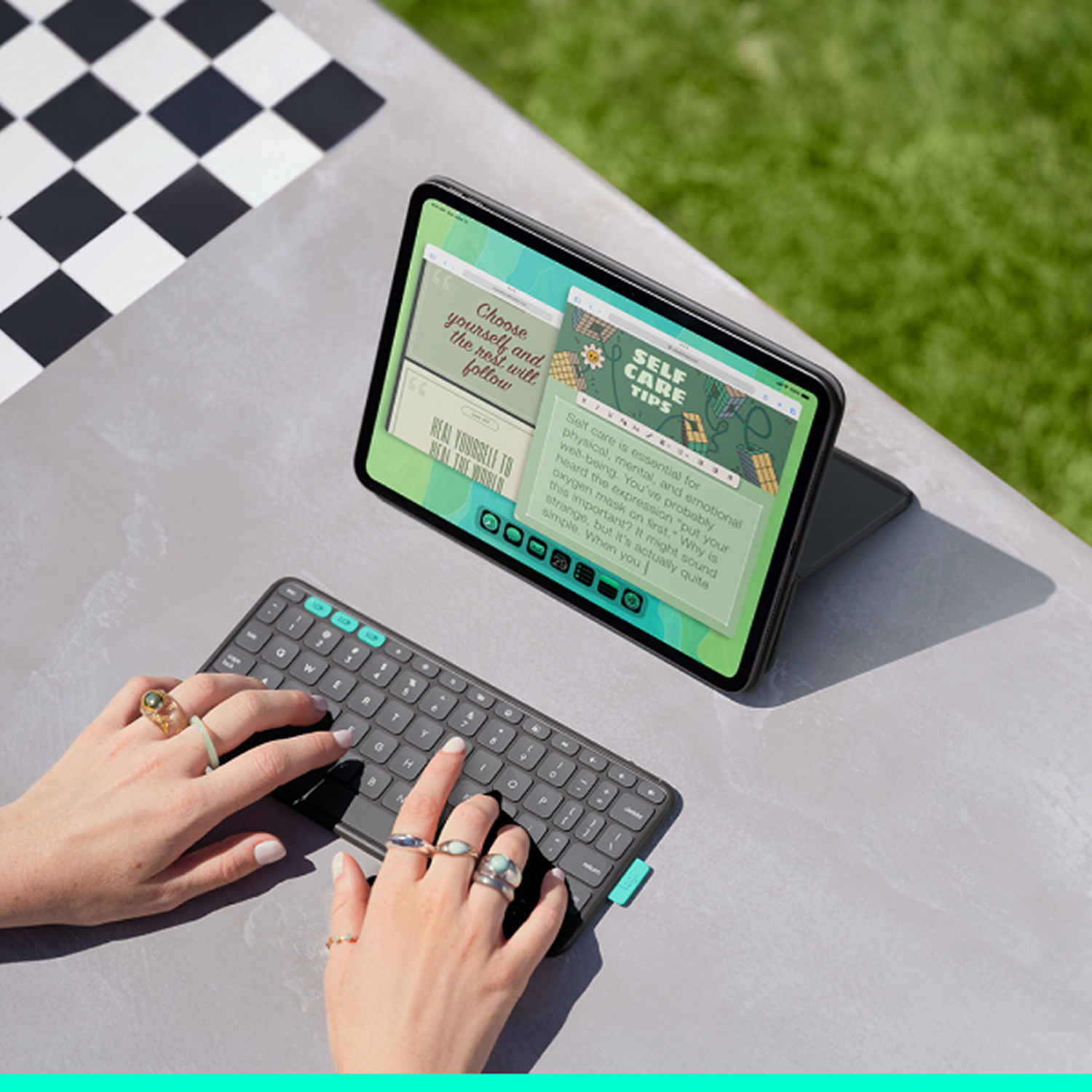 Manos escribiendo en un teclado, una tableta mostrando consejos de autocuidado, anillos en los dedos, sobre una mesa con patrón de cuadros.