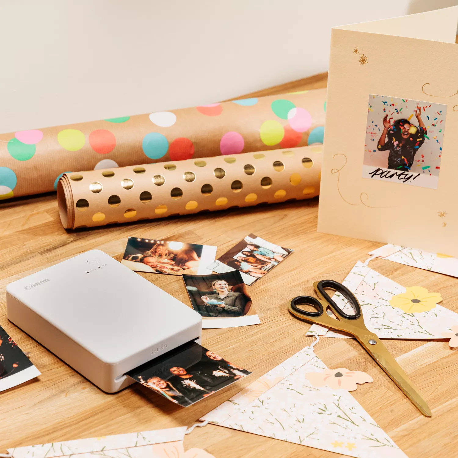Drukarka drukuje zdjęcia na drewnianym stole. Są papiery do pakowania prezentów, karta ze zdjęciem, nożyczki i dekoracyjne flagi.