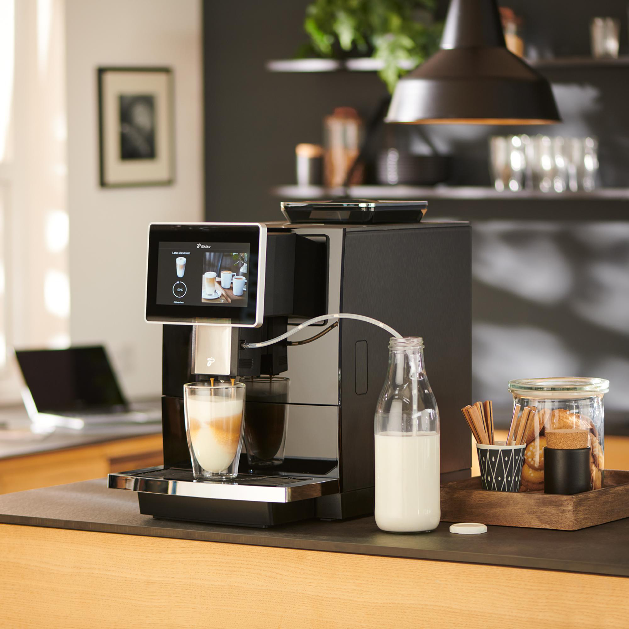 Eine schwarze Kaffeemaschine bereitet einen Latte Macchiato zu, mit Milch und Keksen auf der Theke.