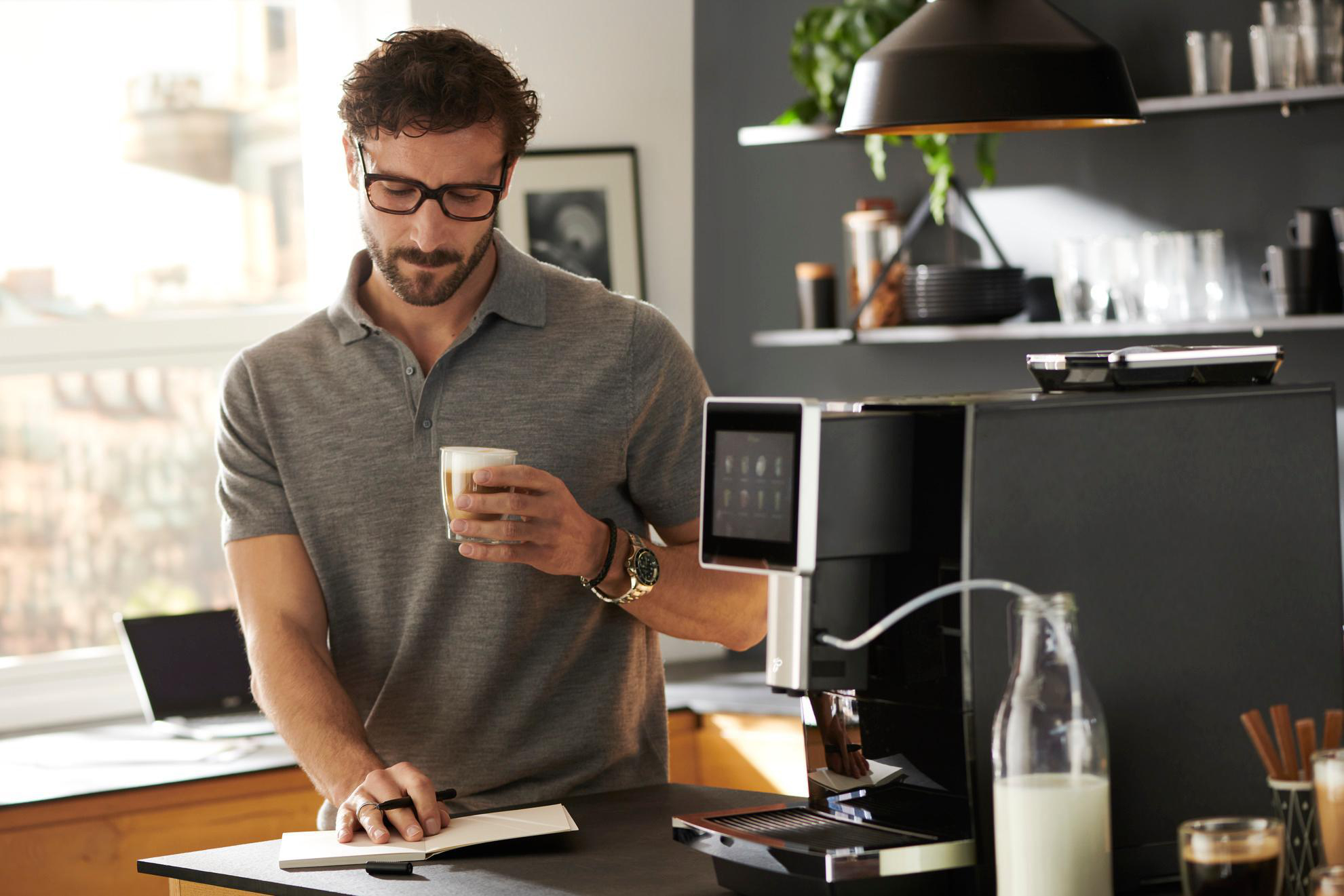 Mann mit Brille hält Kaffee, in der Nähe einer Kaffeemaschine. Notizbuch und Laptop auf dem Tisch. Küchenhintergrund.