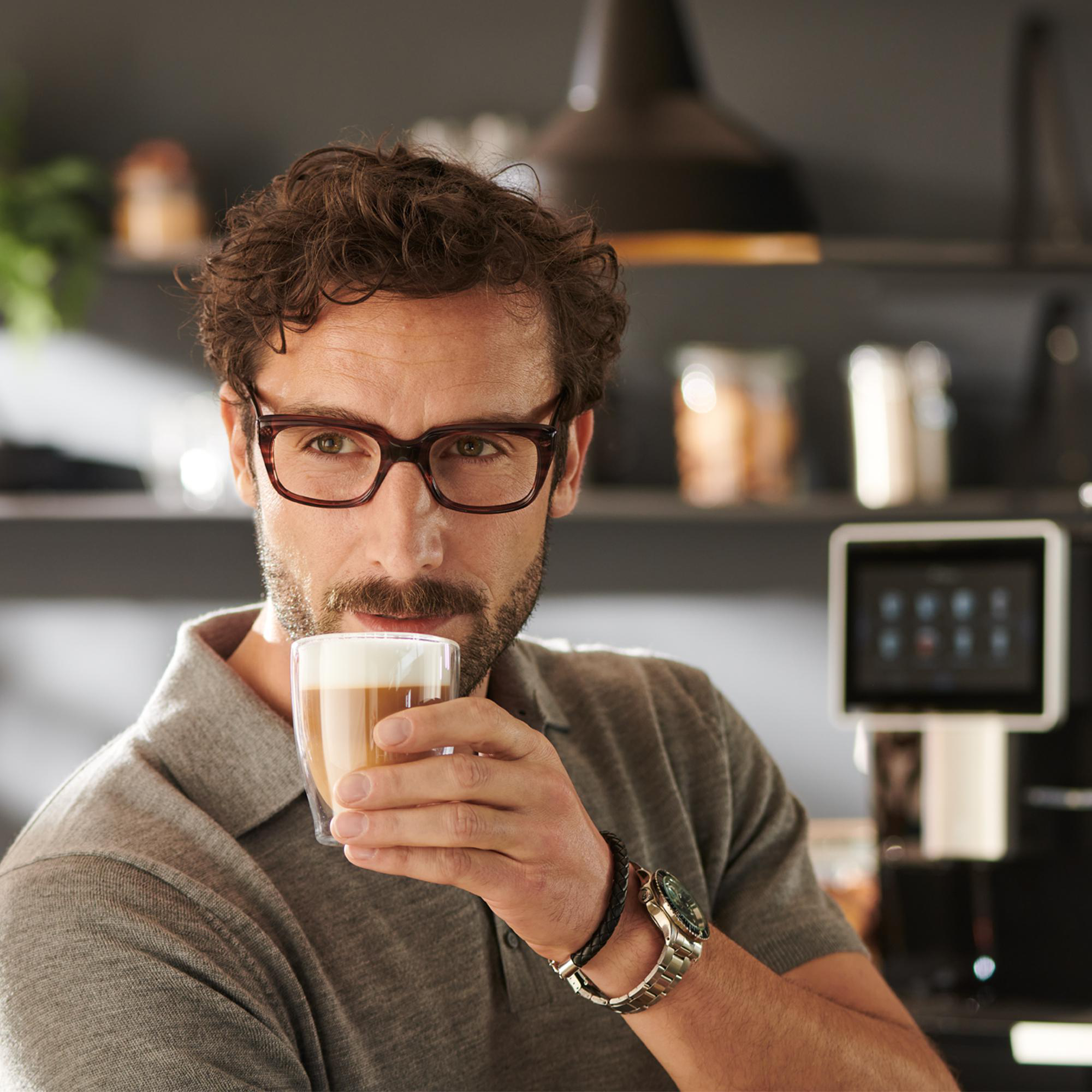 Ein Mann mit Brille trinkt Kaffee. Eine Kaffeemaschine ist im Hintergrund.
