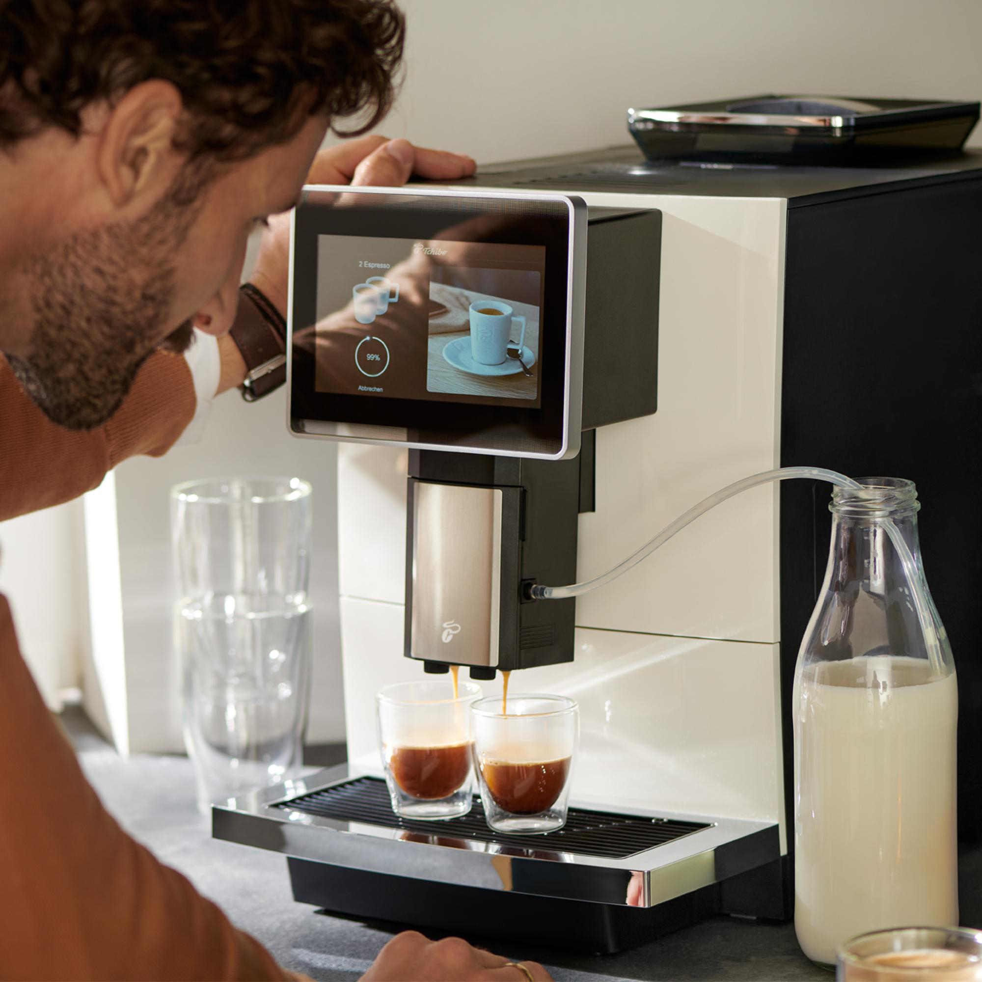 Ein Mann beobachtet, wie Espresso aus einer Kaffeemaschine zubereitet wird. Zwei Gläser werden gefüllt. Rechts steht eine Milchflasche.