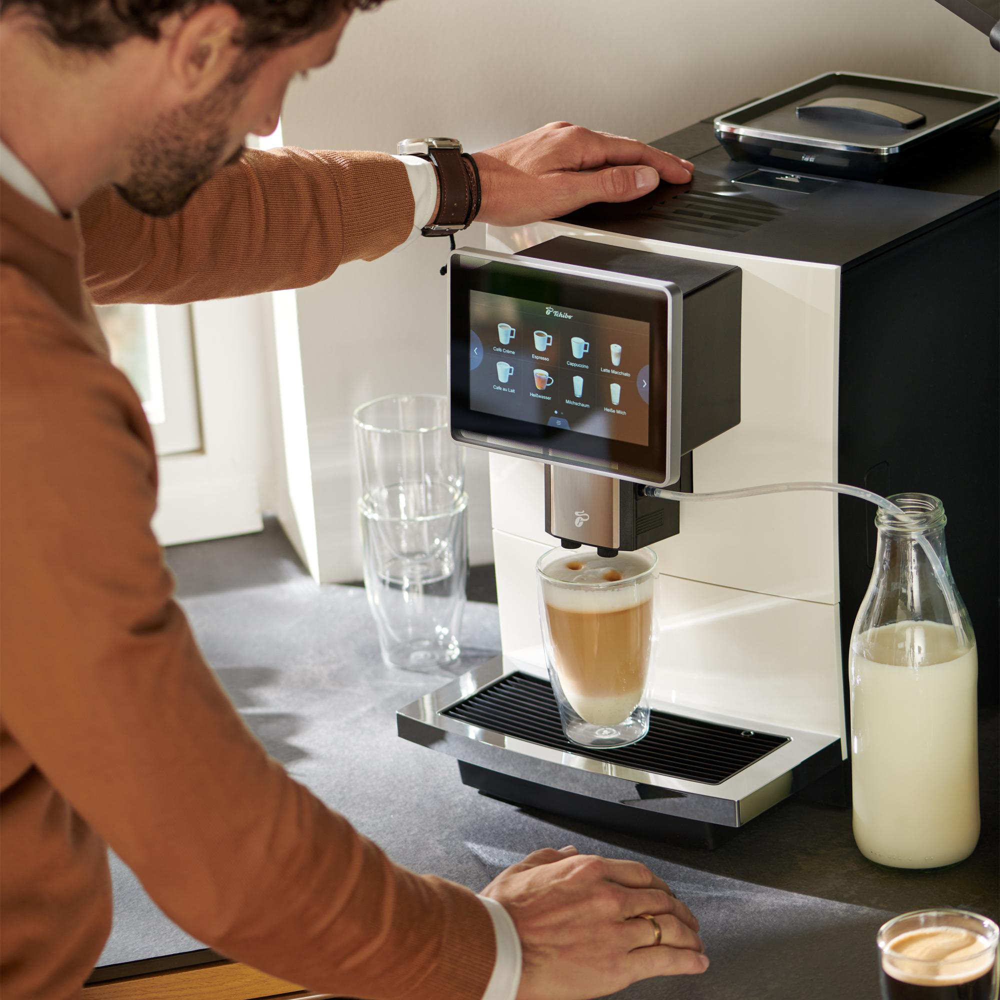Ein Mann bereitet Kaffee zu. Ein Glas Latte Macchiato steht unter der Maschine. Milchflasche in der Nähe.