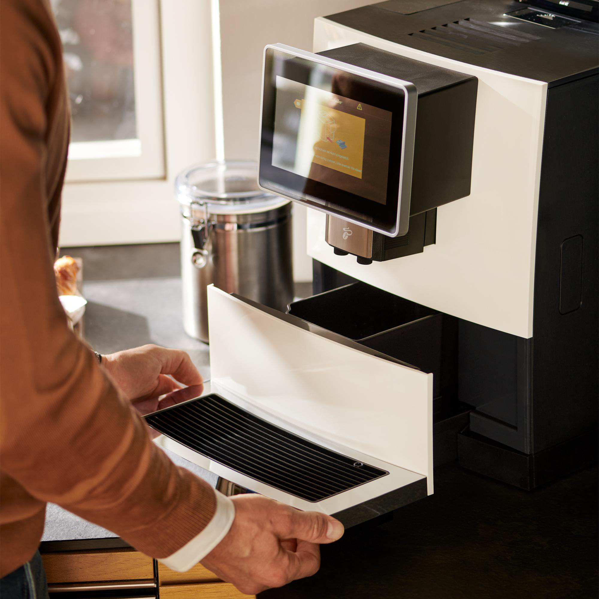 Eine Person setzt ein Tablett in eine Kaffeemaschine mit Touchscreen ein. Die Maschine ist schwarz und weiß.