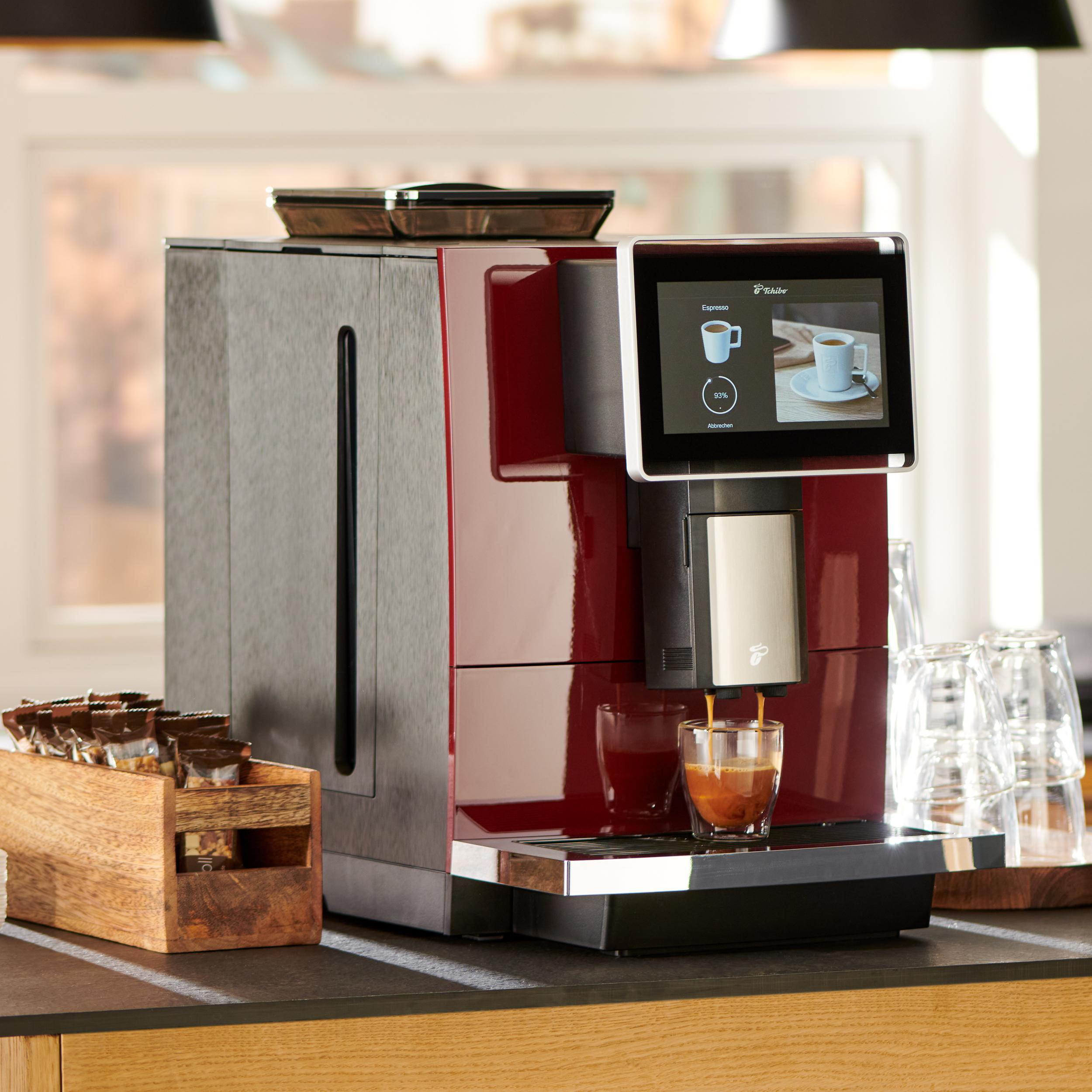 Eine rot-graue Kaffeemaschine, die Espresso in ein Glas ausgibt. Holzbox mit Snacks links. Hintergrund beinhaltet Fenster.