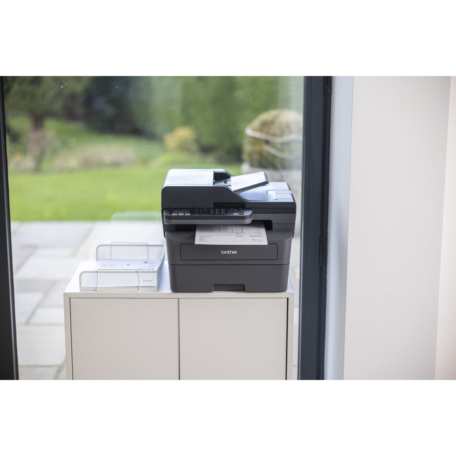 A black printer on a white cabinet. A document is printing. Window shows a garden.