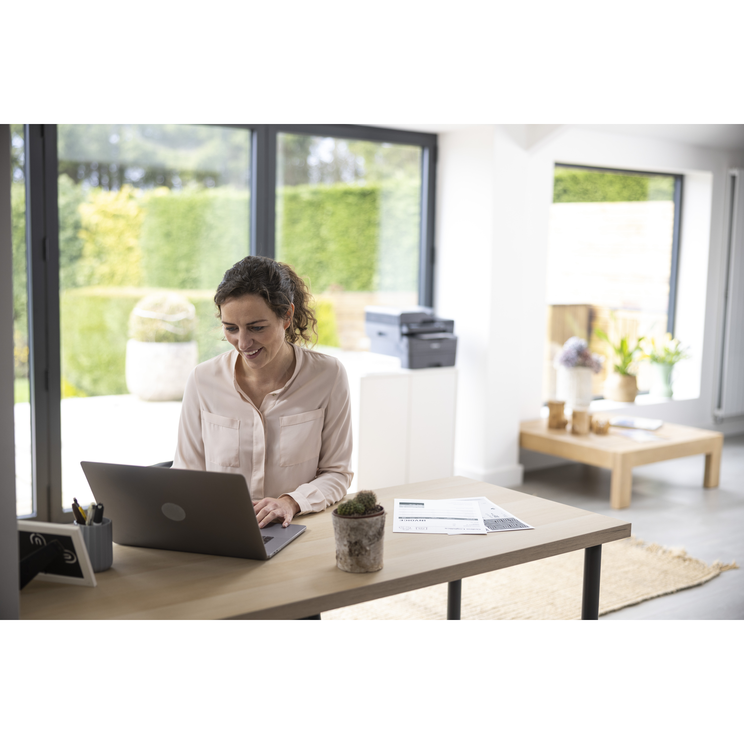 Woman working on laptop at desk. Cactus, printer, and papers are visible. Background features a garden and wooden table.