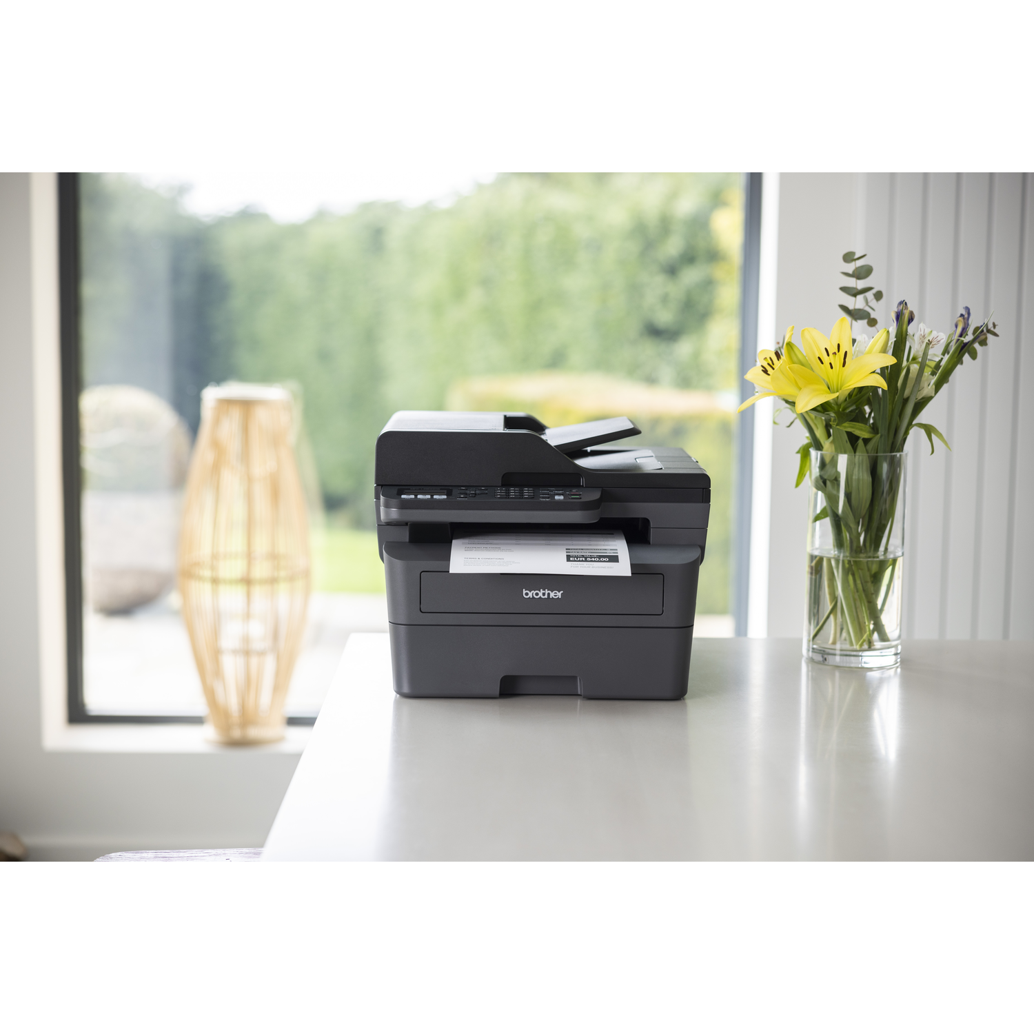 Black Brother printer on a white table, with flowers in a vase to the right. A wicker decoration is on the left.