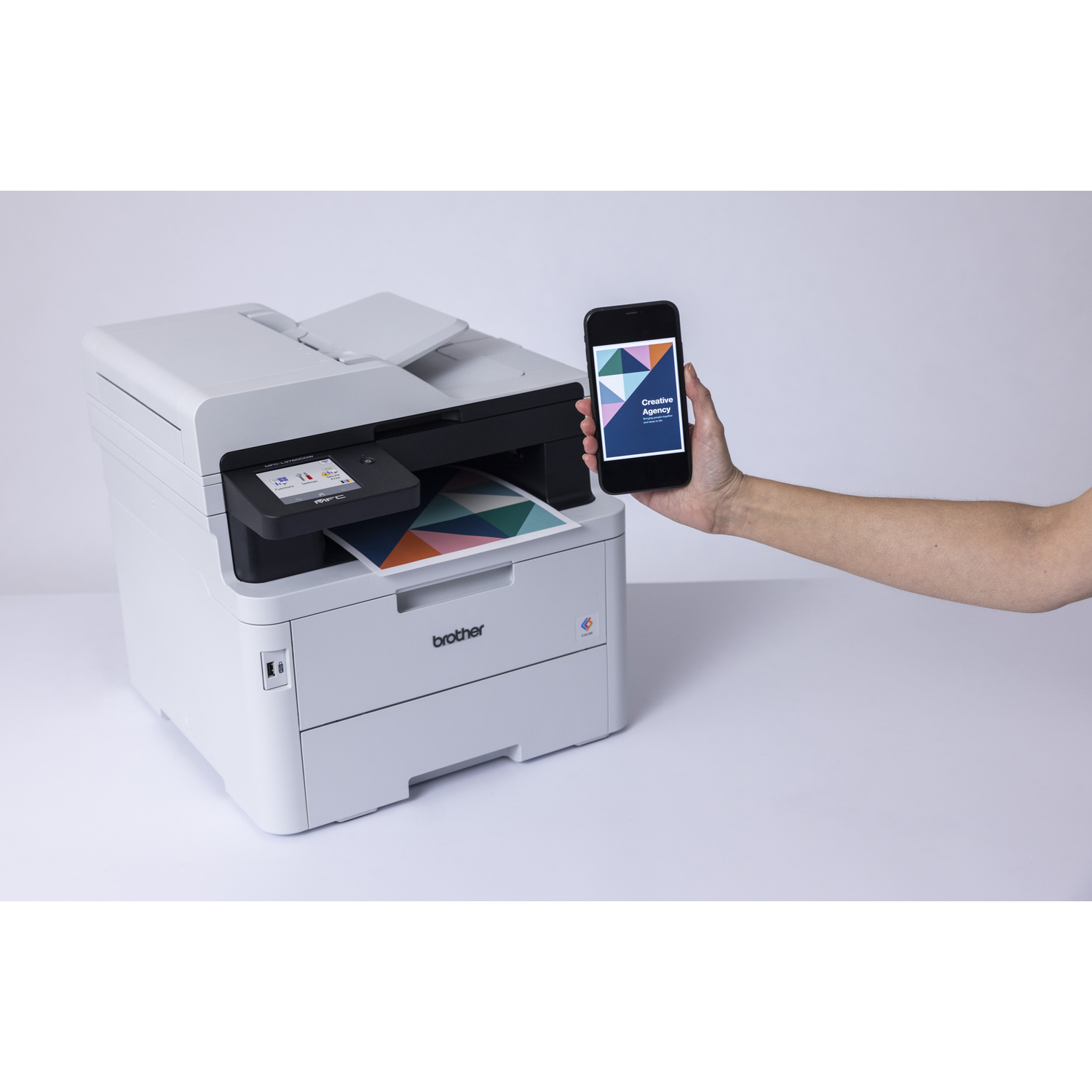 Een witte Brother-printer met papieruitvoer, naast een hand die een telefoon vasthoudt. Telefoon toont een kleurrijk ontwerp.