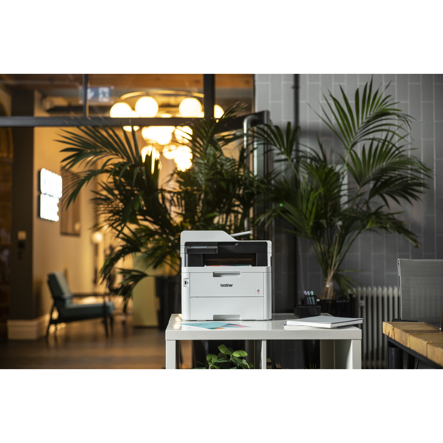Een witte Brother printer op een witte tafel met een plant. Op de achtergrond een kamer met stoel en warme verlichting.