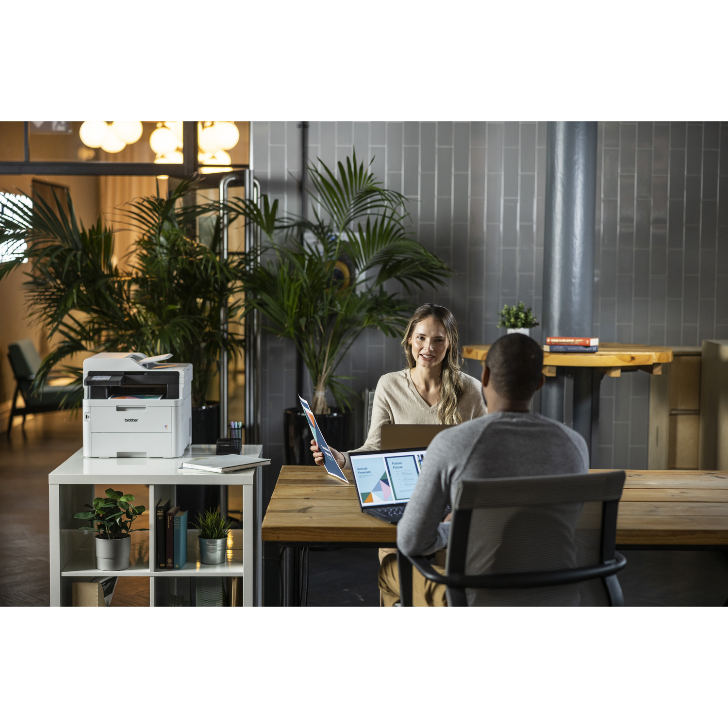 Twee mensen aan een tafel, een toont een document. Een printer en planten op de achtergrond. Moderne kantooromgeving.