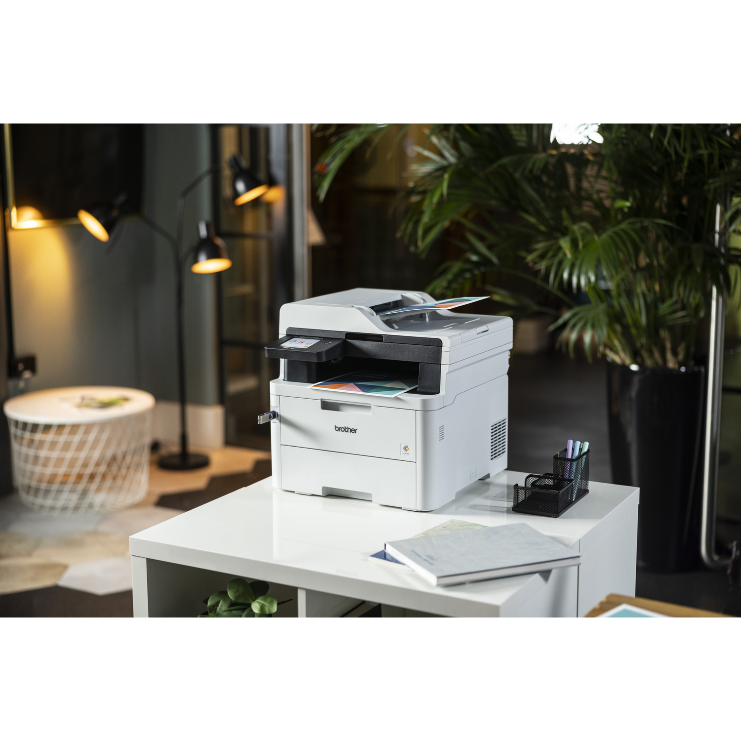 Witte printer op een witte tafel. Er staat een plant op de achtergrond en een lamp. Schrijfwaren op de tafel.
