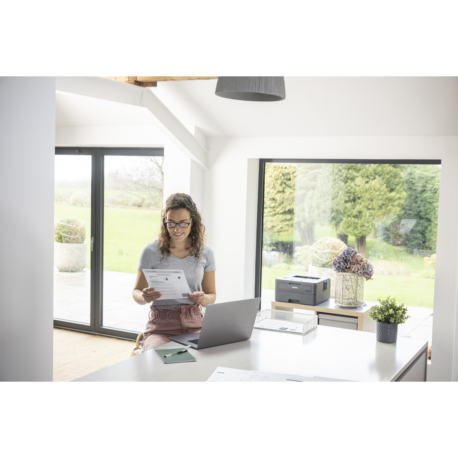 Vrouw met bril leest document bij laptop. Printer en planten op achtergrond bij raam.