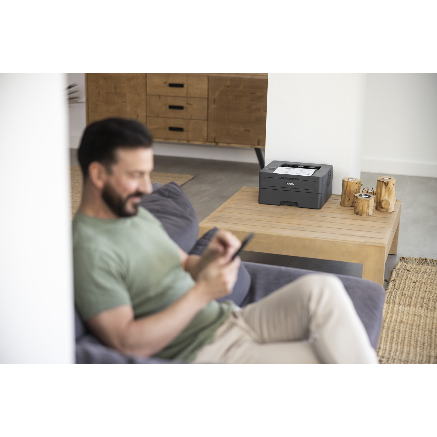 Man gebruikt telefoon, kijkt naar zwarte printer op tafel. Houten decor en kast op de achtergrond.