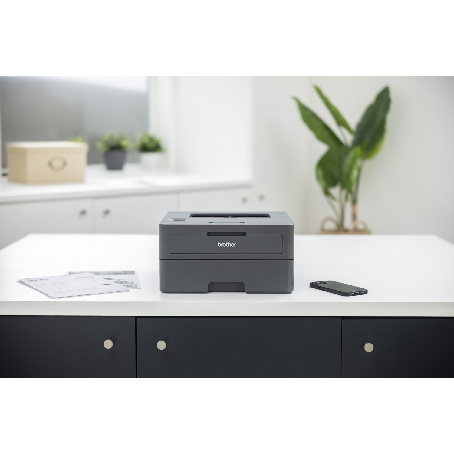Een Brother-printer staat op een witte tafel met een telefoon en papieren. Een doos en planten staan op de achtergrond.