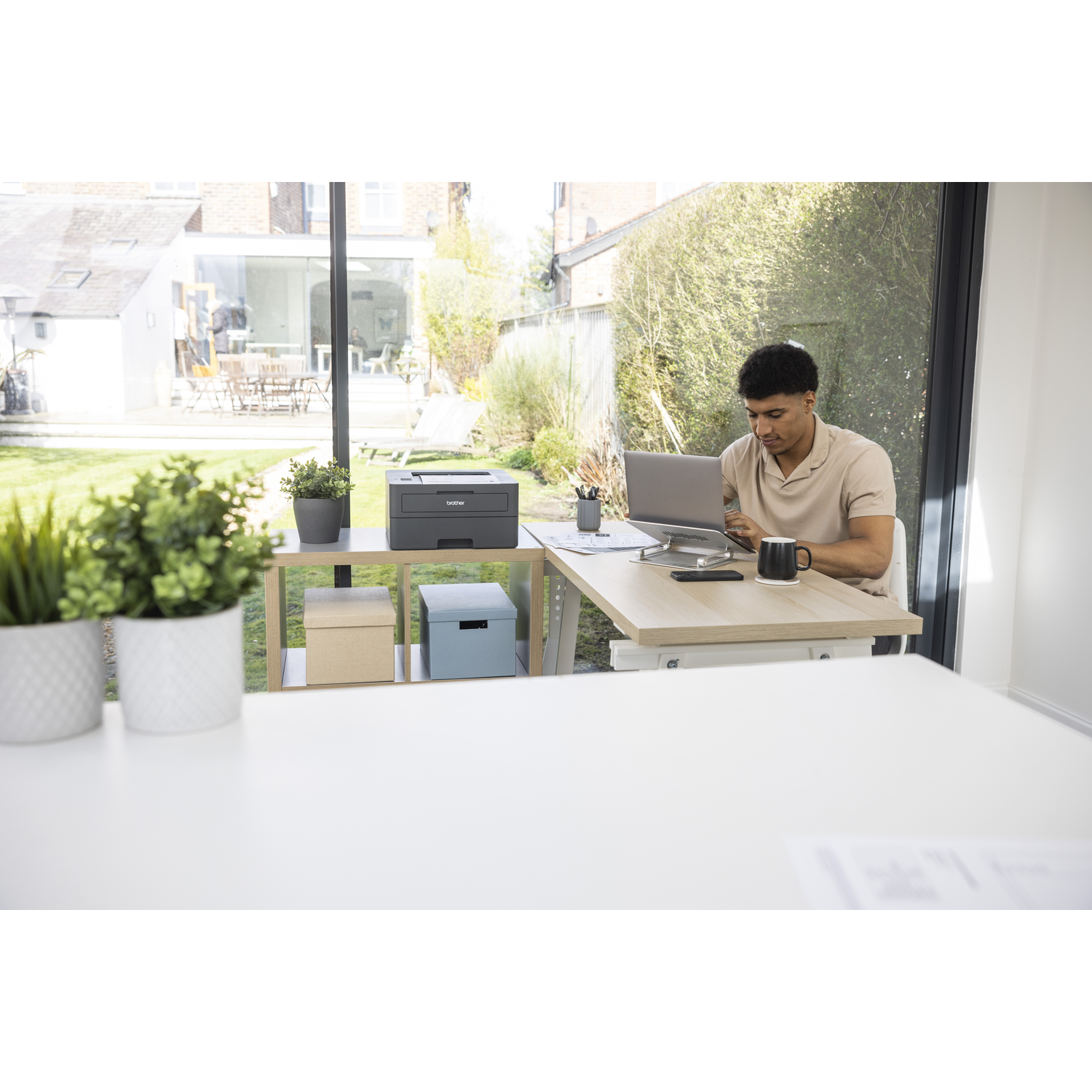 Man werkt op laptop aan bureau. Printer en dozen op plank. Achtertuin zichtbaar door raam.
