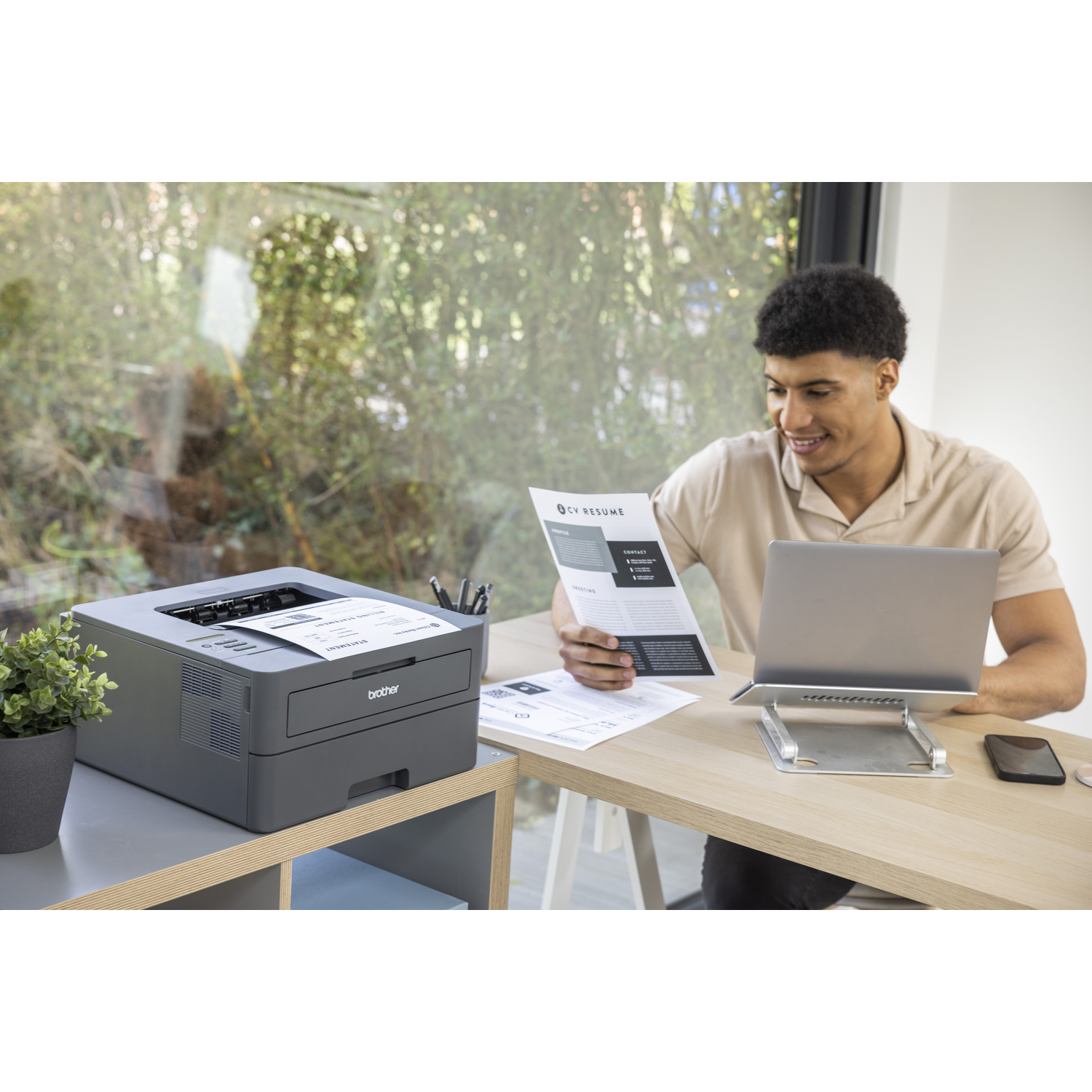 Man bekijkt cv naast laptop. Printer en plant links. Achtergrond toont raam met bomen.