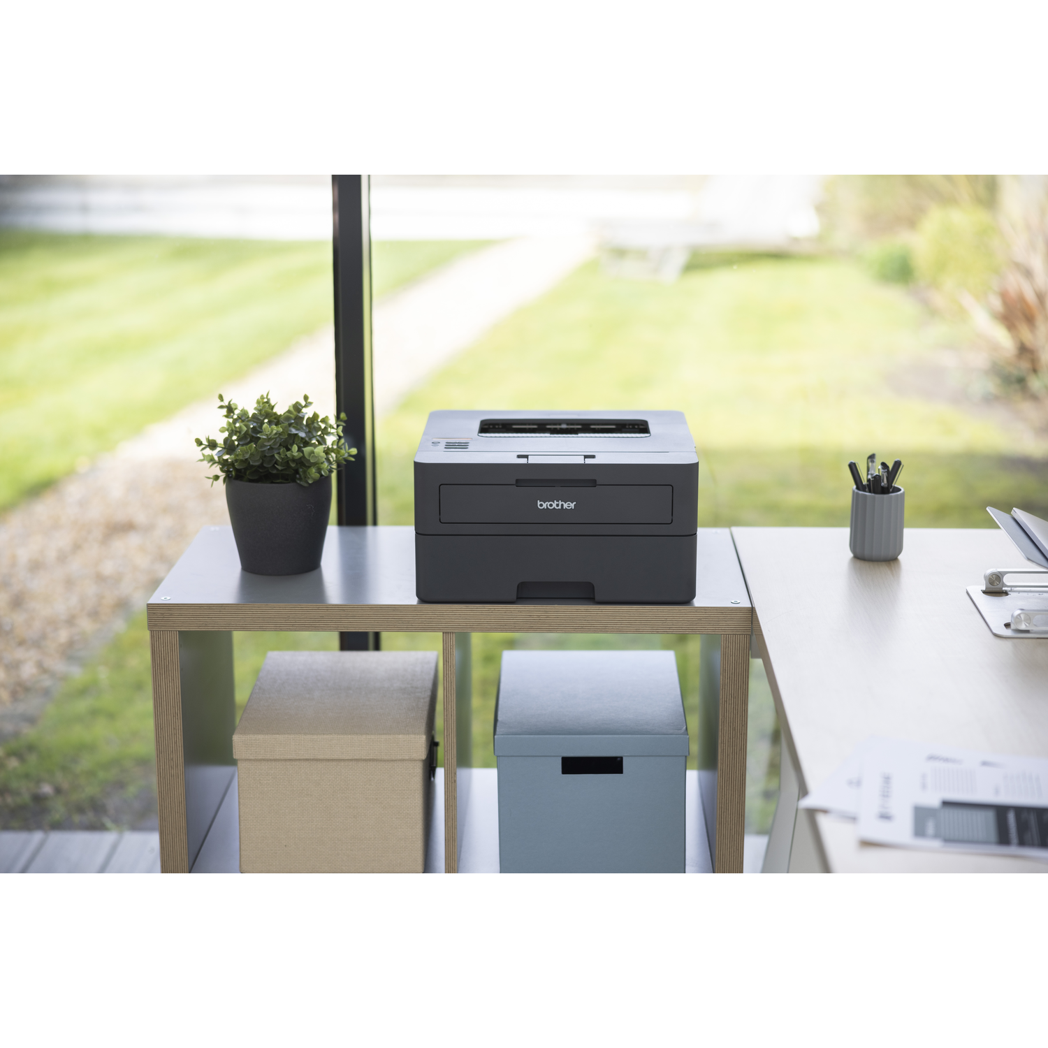 Een Brother-printer staat op een bureau met een plant, dozen en pennen. Het raam op de achtergrond toont een groen gazon.