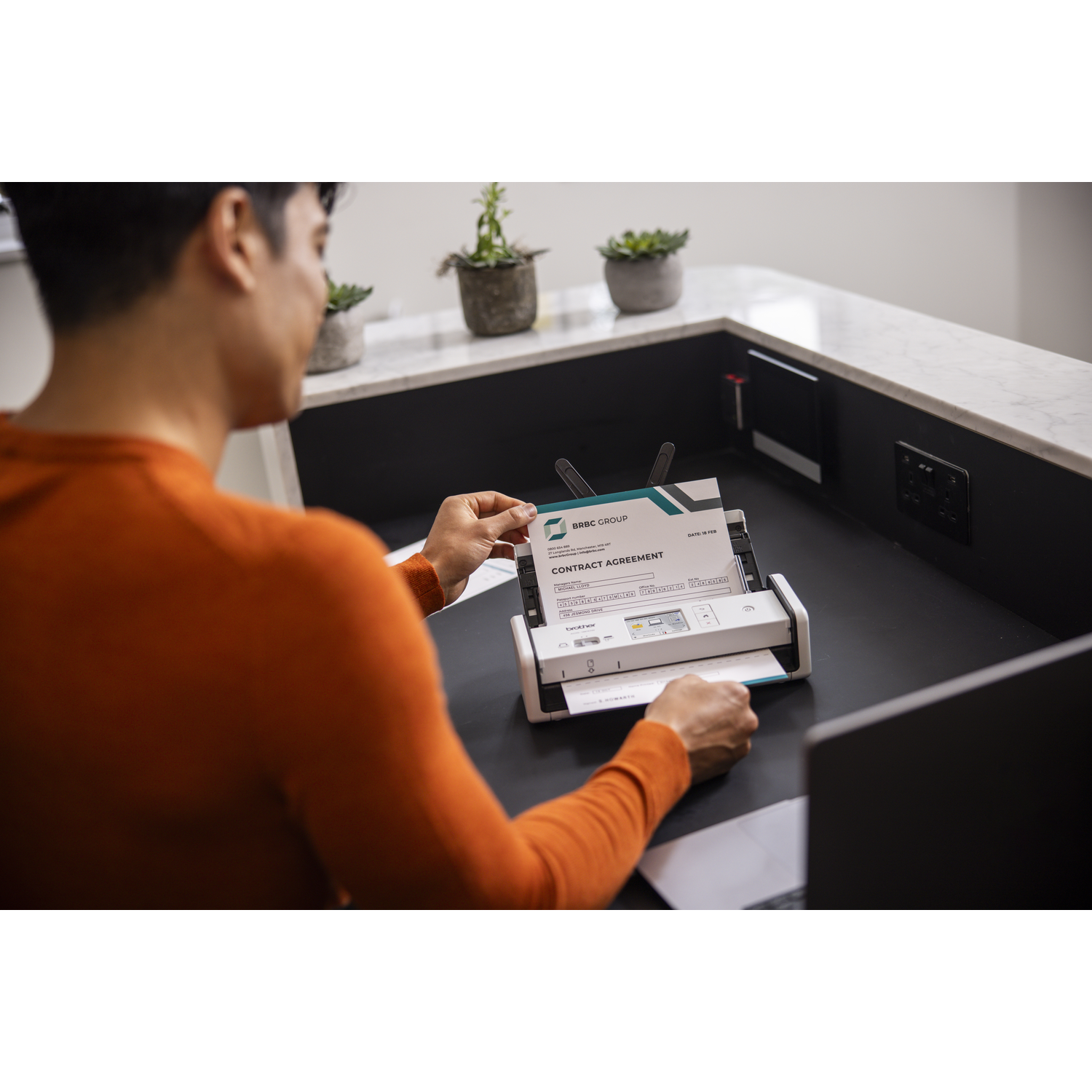 Een persoon scant een contractovereenkomst. Er staat een scanner en een laptop op een zwart bureau. Potten met planten op de achtergrond.