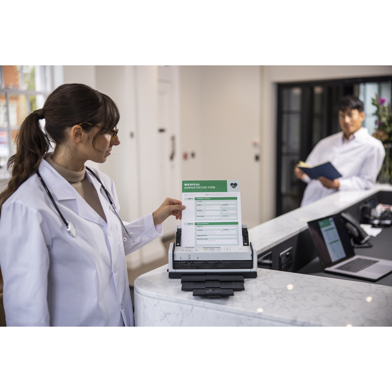 Een dokter scant een medisch formulier. Een andere persoon op de achtergrond houdt een boek vast. Een laptop en andere spullen staan op een bureau.