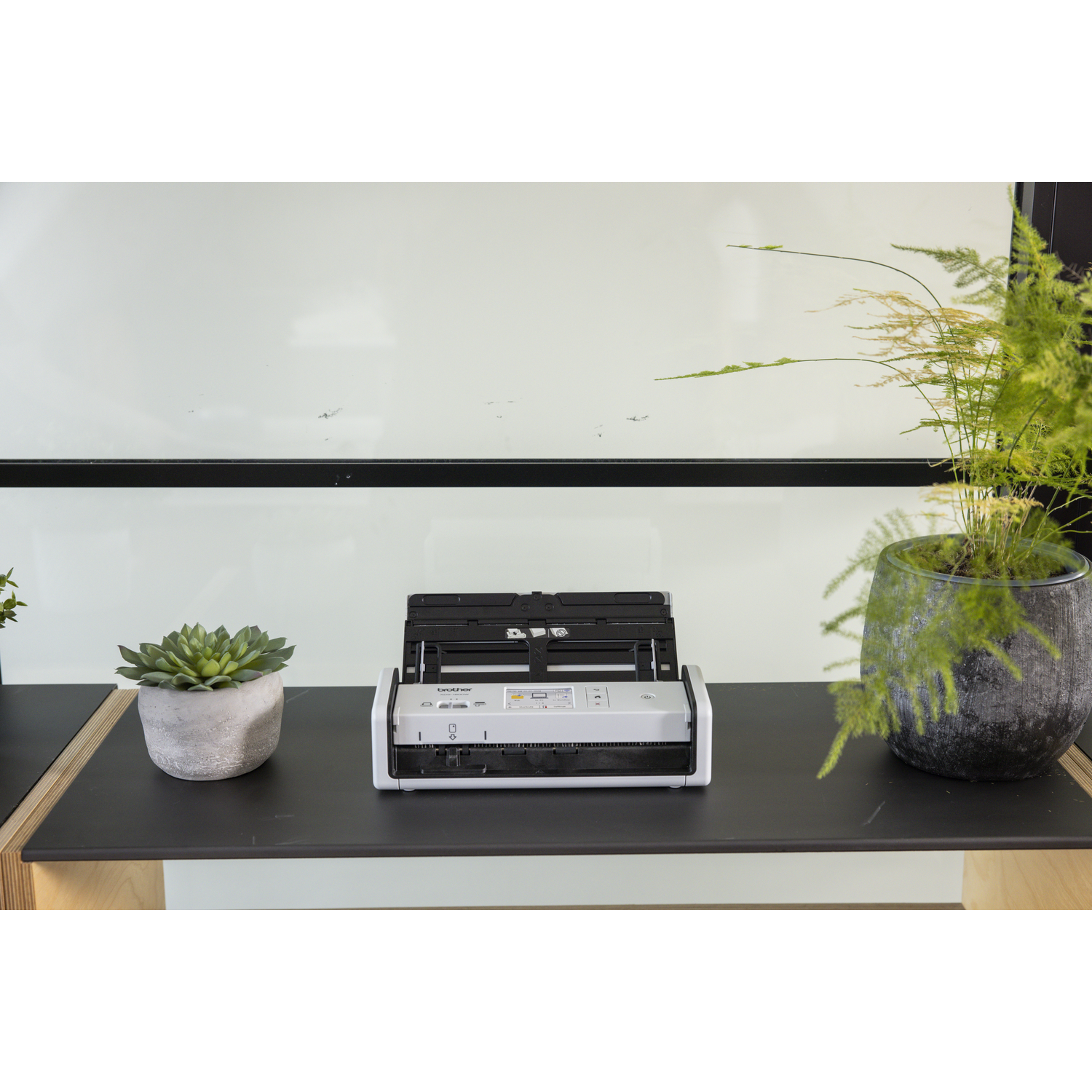 Een open scanner op een zwarte plank, met een vetplant en een varen. Achtergrond: een witte muur met een zwarte rand.
