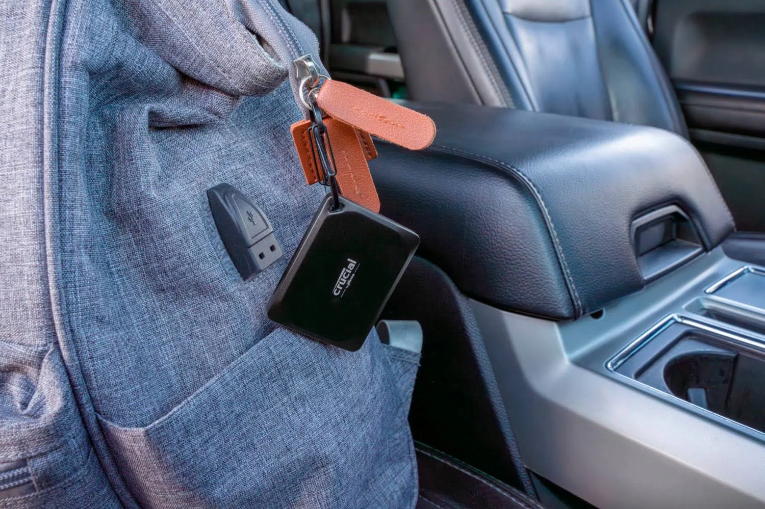 Una mochila gris con una unidad USB y llaves. Una unidad Crucial negra cuelga de las llaves. Interior del coche en el fondo.