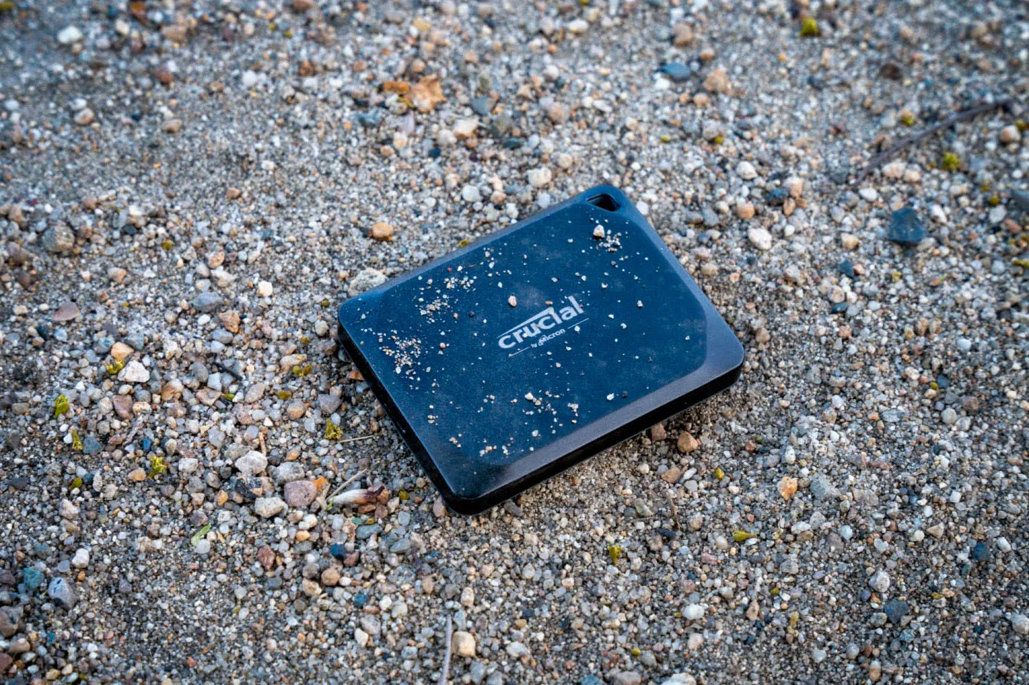Un disco duro externo Crucial negro sobre grava y piedras pequeñas. La escena es al aire libre, enfocada en detalles.