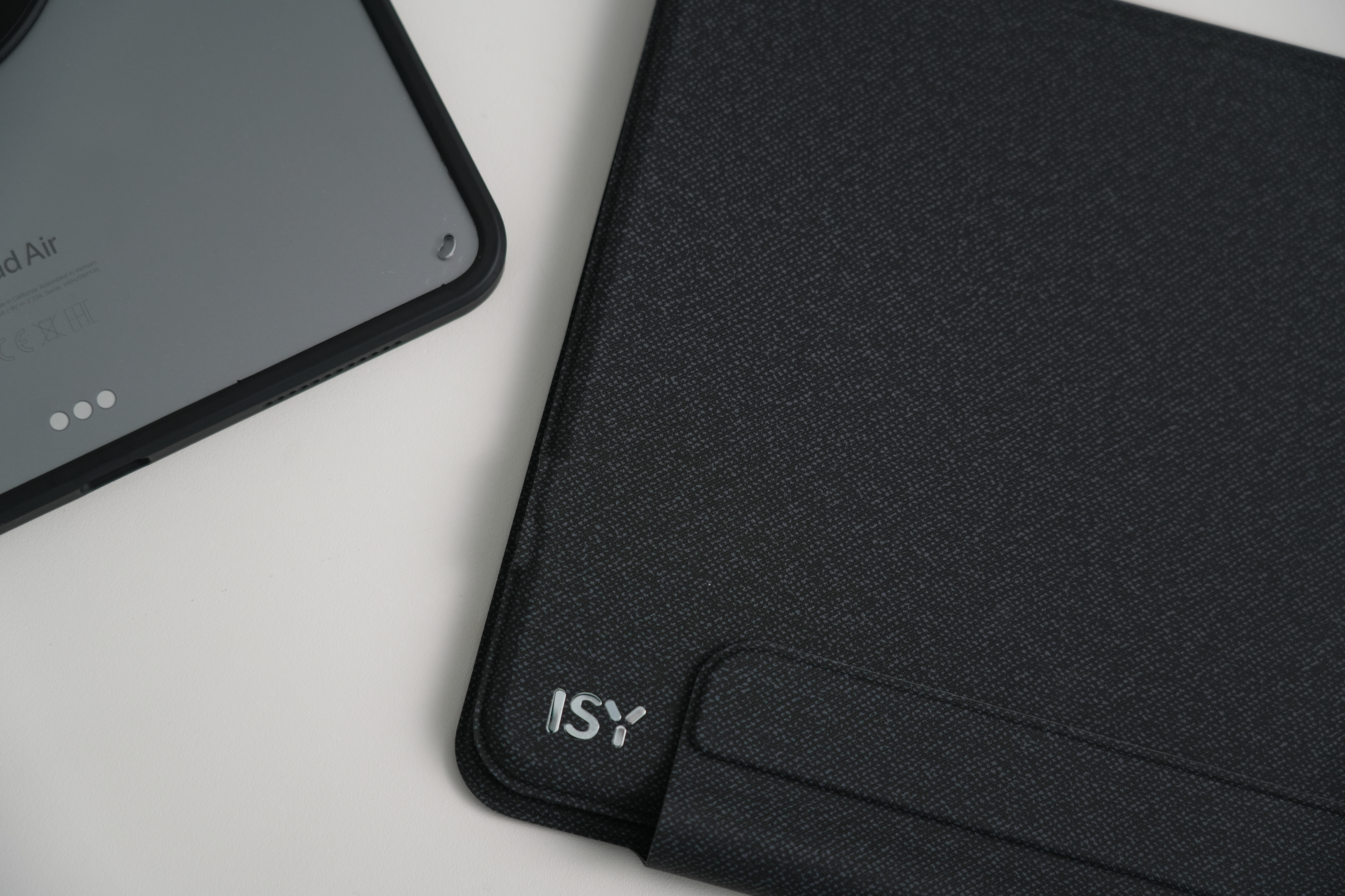 Ein graues Tablet und eine schwarze gemusterte Hülle liegen auf einer weißen Oberfläche. Auf der Hülle steht ISY.