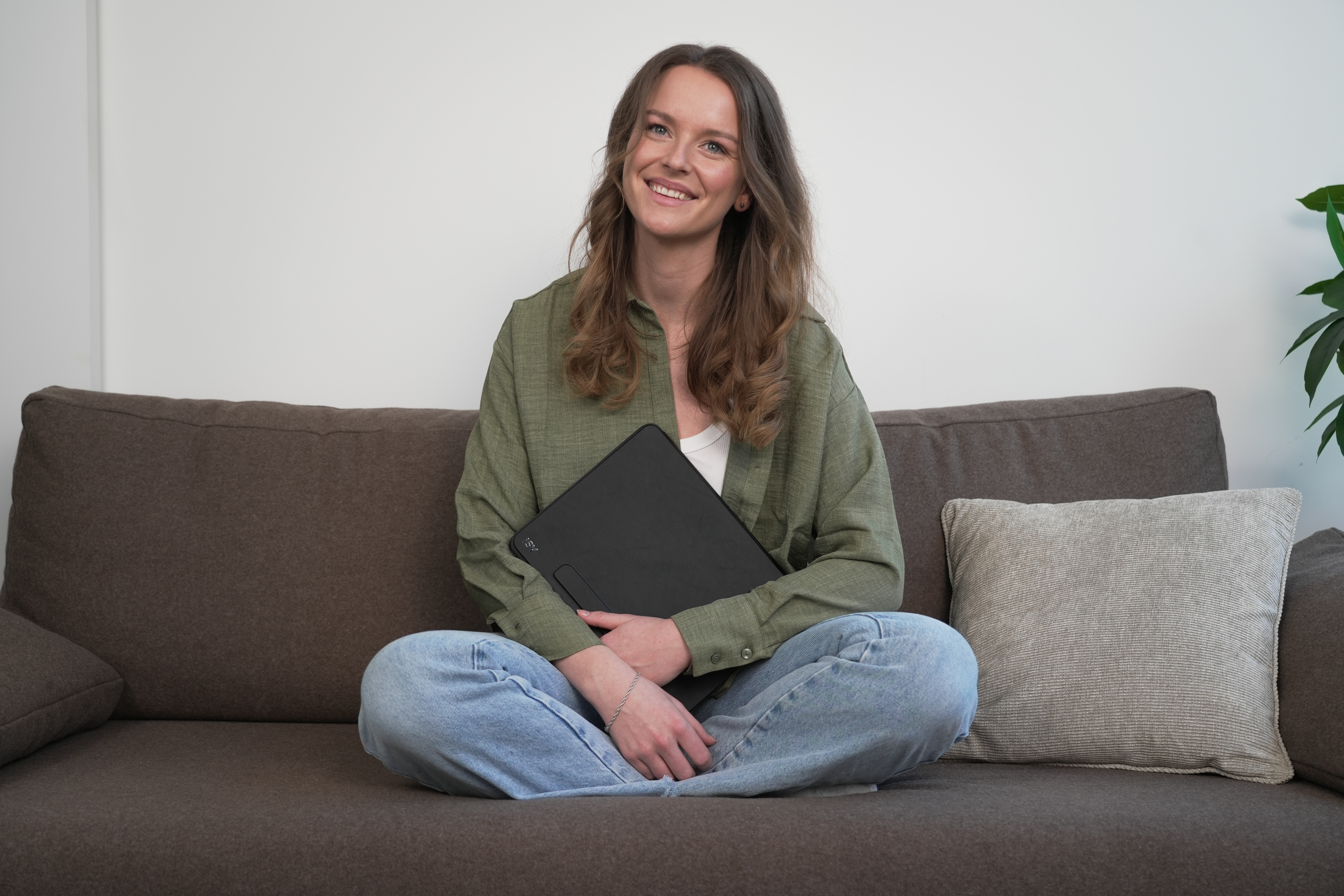 Frau sitzt auf Couch, Tablet haltend. Sie trägt grünes Hemd und Jeans, lächelt. Braune Couch, weiße Wand.