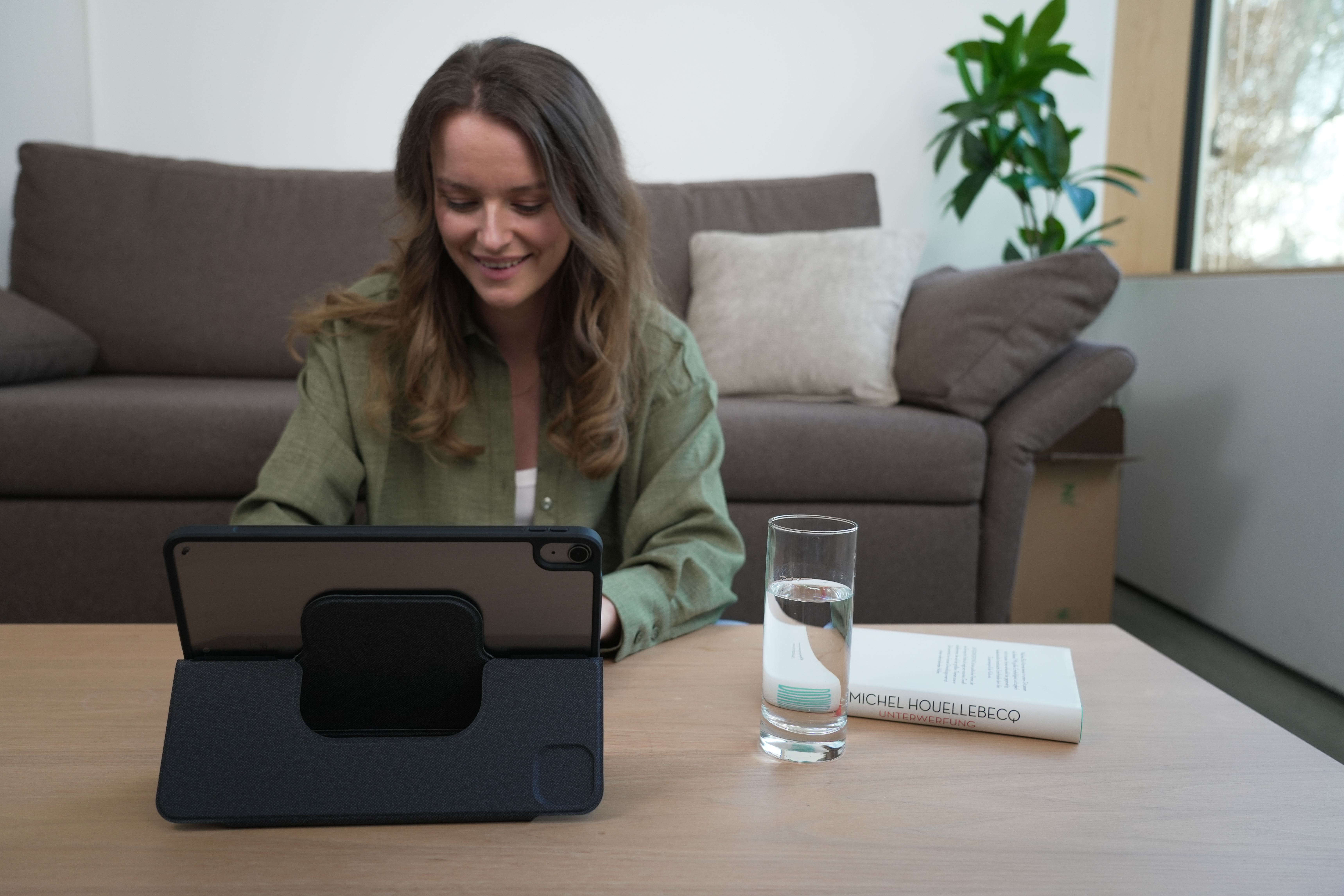 Frau benutzt Tablet, lächelt, neben Glas Wasser und Buch. Braunes Sofa im Hintergrund.