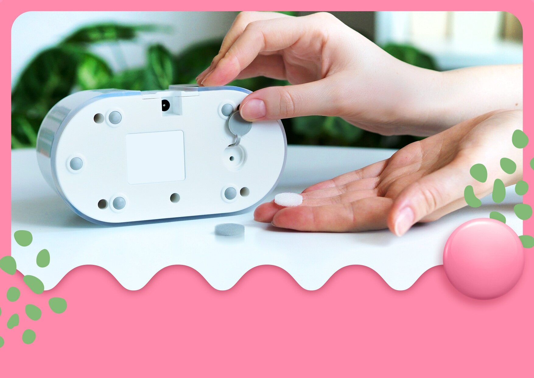 Dłonie umieszczają białą, okrągłą podkładkę na białym urządzeniu. Kolejna podkładka leży na stole. Zielone rośliny są rozmyte w tle.