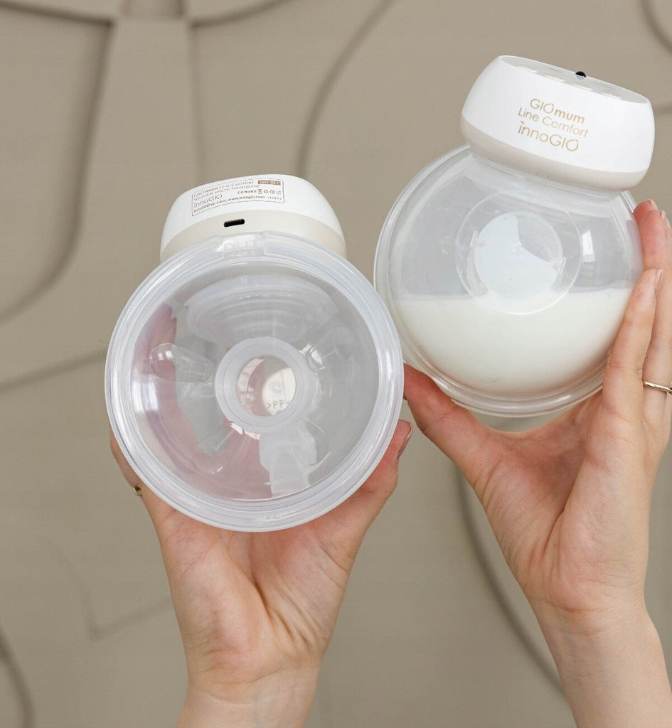 Dwie dłonie trzymają laktatory. Jeden ma mleko. Oba są przezroczyste z białymi nakładkami, na beżowym tle.