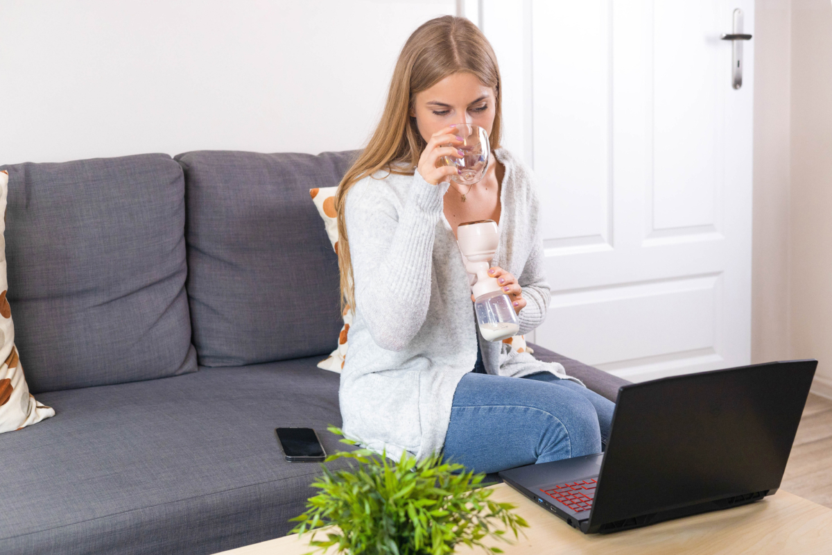 Kobieta na kanapie pije wodę, trzyma laktator, laptop i roślinę na stole.