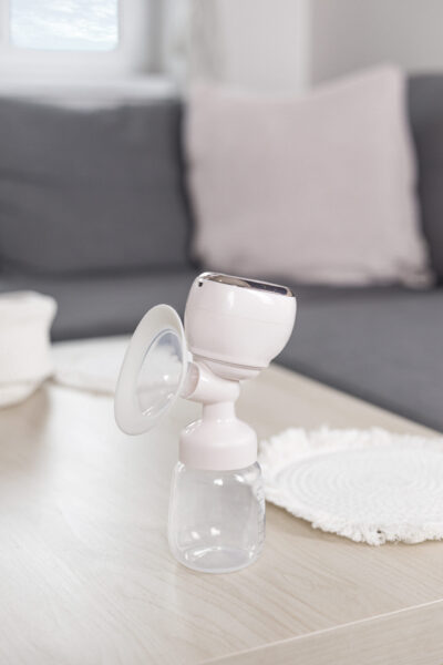 Biało-różowa laktator stoi na jasnym drewnianym stole. Szara sofa i różowa poduszka są w tle.