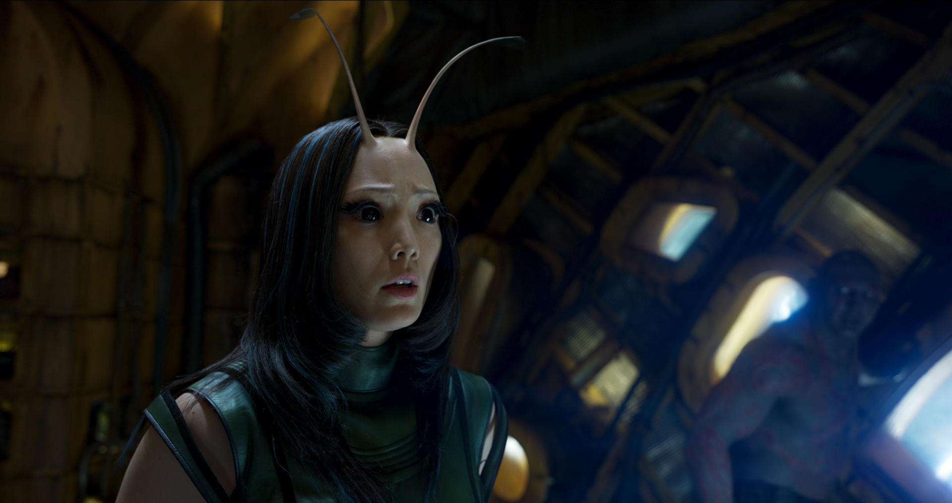 Mantis mit Antennen sieht besorgt aus. Drax ist im Hintergrund. Innenraum eines Raumschiffs. Dunkel, mit Grün- und Gelbtönen.
