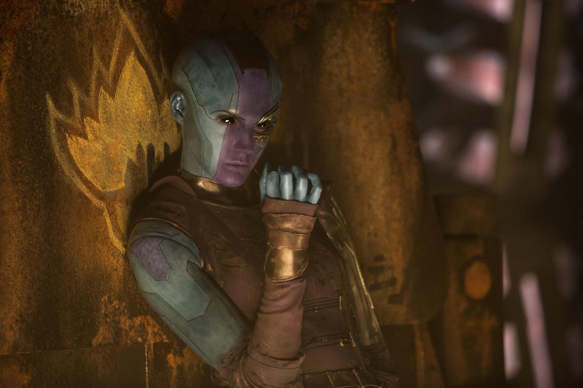 Nebula, ein Cyborg mit blauer Haut, lehnt an einer Wand. Sie hat einen strengen Ausdruck und trägt ein dunkles Outfit mit goldenen Akzenten. Der Hintergrund ist dunkel.