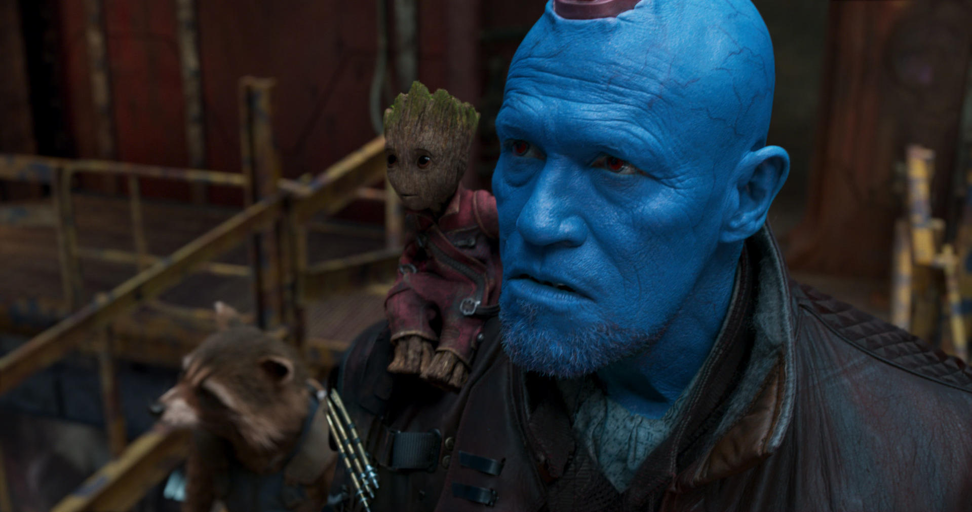 Yondu mit Groot auf der Schulter und Rocket im Vordergrund. Blaue Haut, rote Augen und ein besorgter Ausdruck. Metallischer Hintergrund.