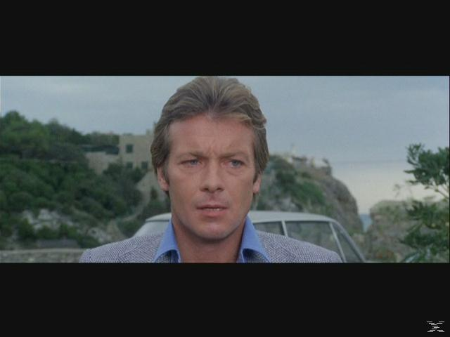 Ein Mann mit blonden Haaren schaut in die Kamera. Hinter ihm sind ein Auto und eine Landschaft mit Bäumen und einem Gebäude. Bewölkter Tag.