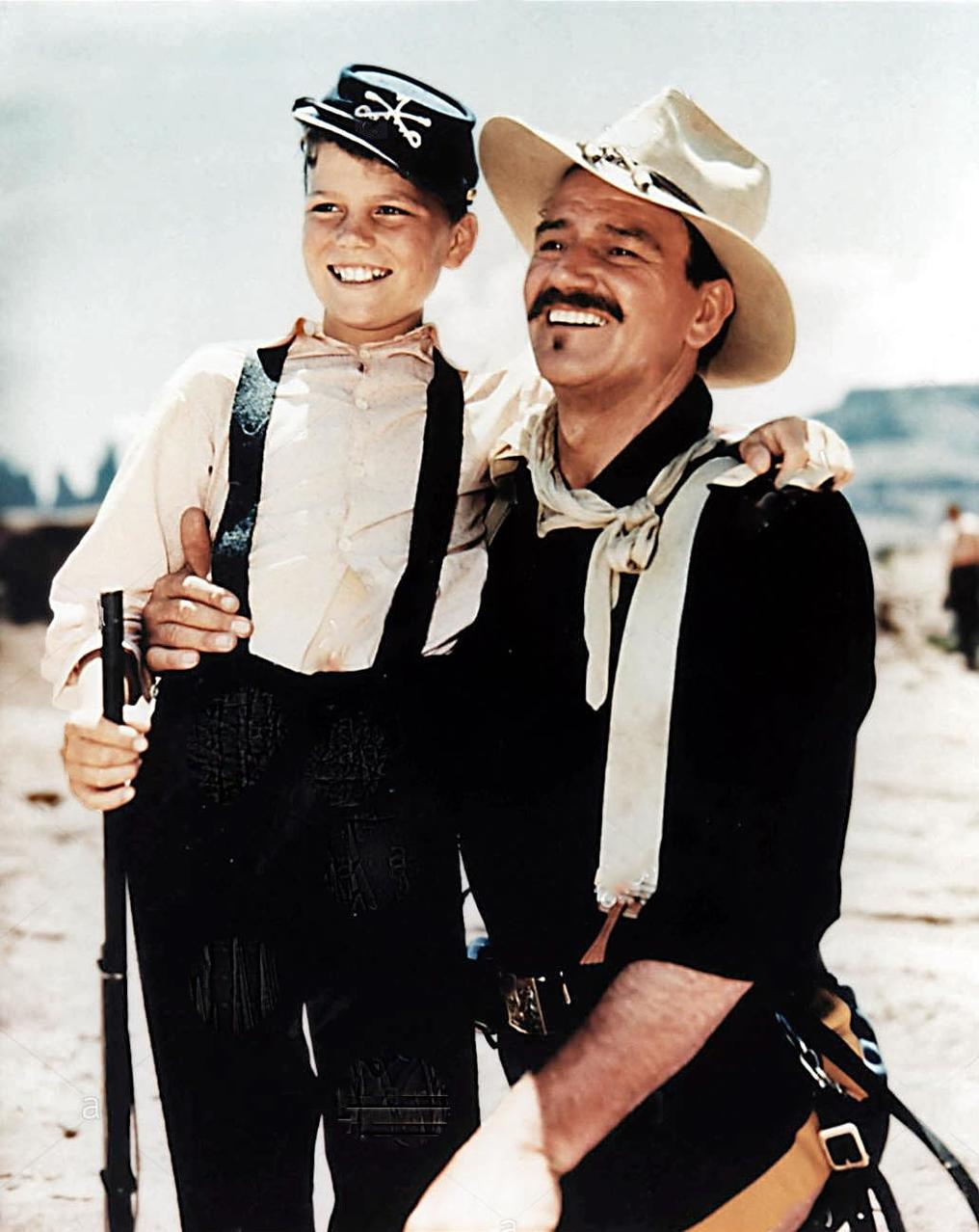 Ein Junge in Uniform und ein Mann mit Cowboyhut lächeln. Der Mann hat einen Schnurrbart, der Junge hält ein Gewehr.