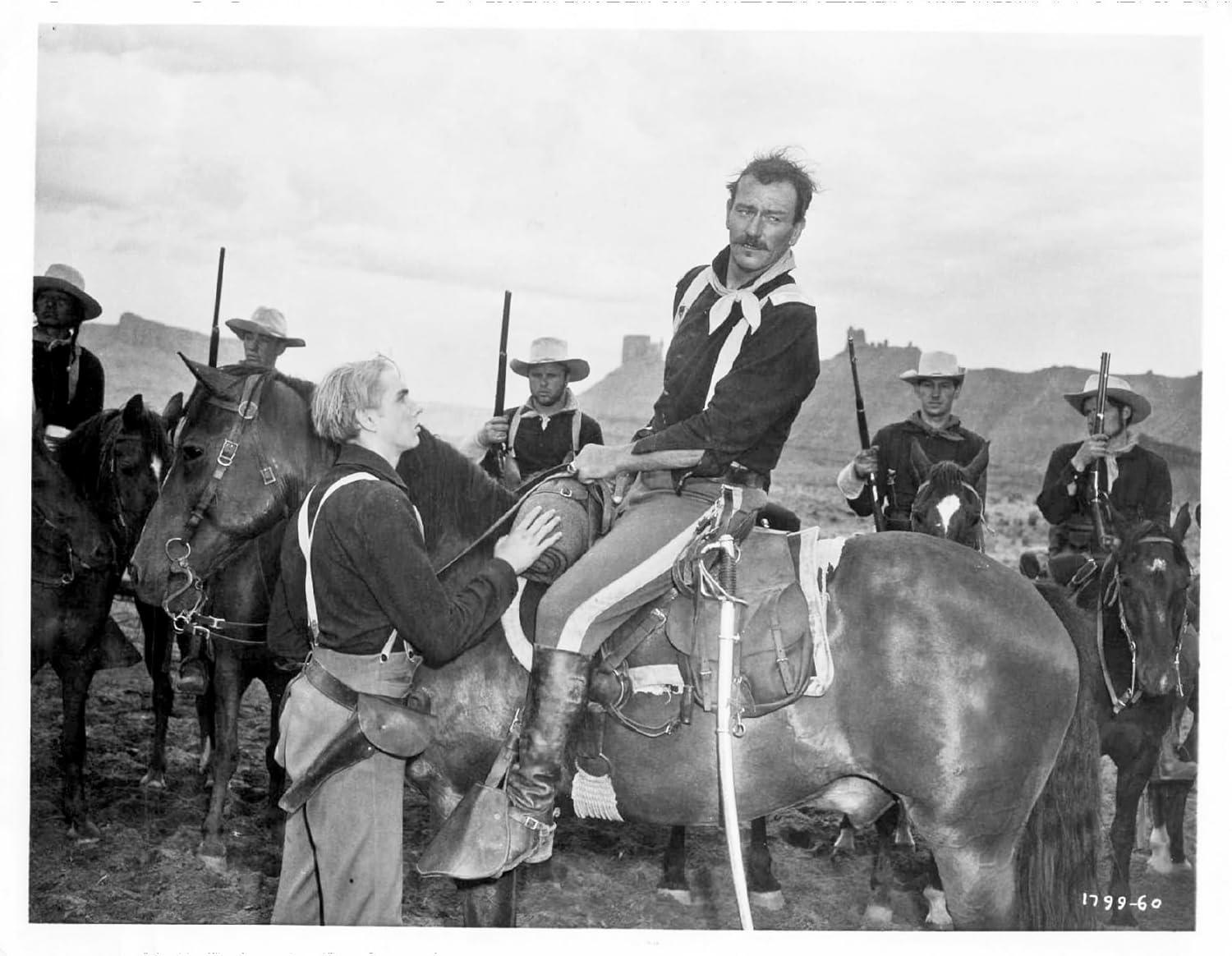 Ein Mann auf einem Pferd wird von einem anderen Mann unterstützt. Andere Männer auf Pferden halten Gewehre. Berge sind im Hintergrund.