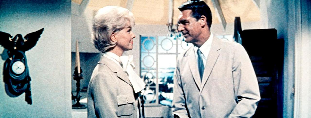 Ein Mann und eine Frau in hellen Anzügen stehen sich gegenüber. Links ist eine Wanduhr zu sehen. Im Hintergrund ist ein Fenster zu sehen.