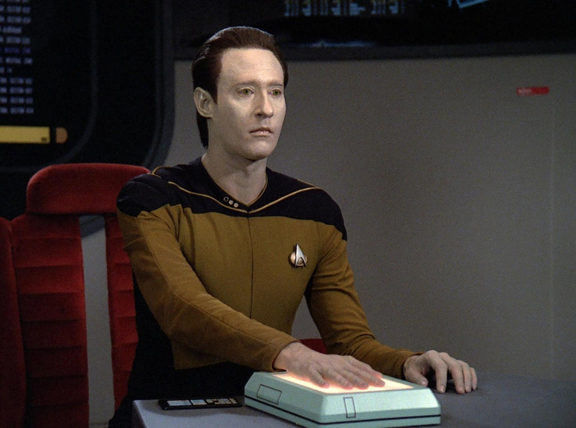 Data aus Star Trek. Blasshäutiger Android in Uniform, berührt ein leuchtendes Panel. Roter Sitz und Bedienfeld im Hintergrund.