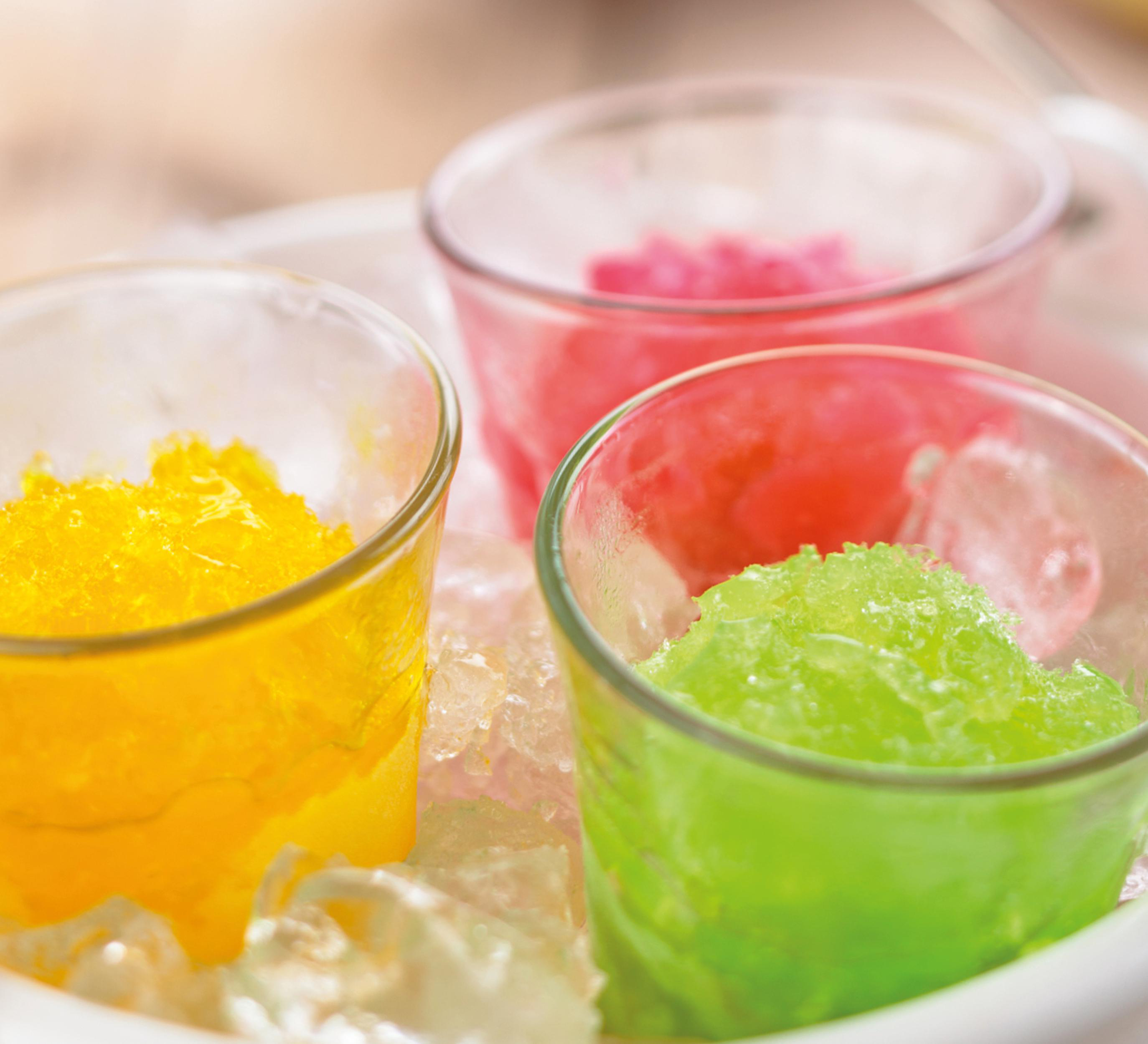 Drei Gläser mit gelbem, rosa und grünem Eisrasur auf einem Bett aus Eiswürfeln.