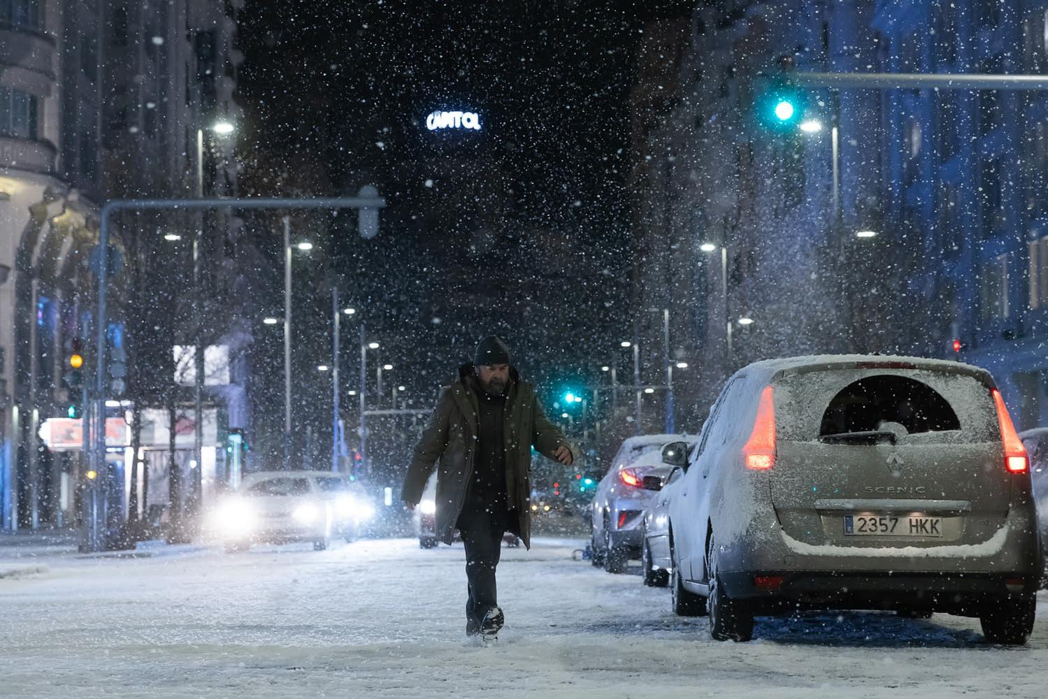 Ein Mann geht nachts durch eine verschneite Straße. Autos parken am Straßenrand. Schnee fällt, und das Wort CAPITOL ist sichtbar.