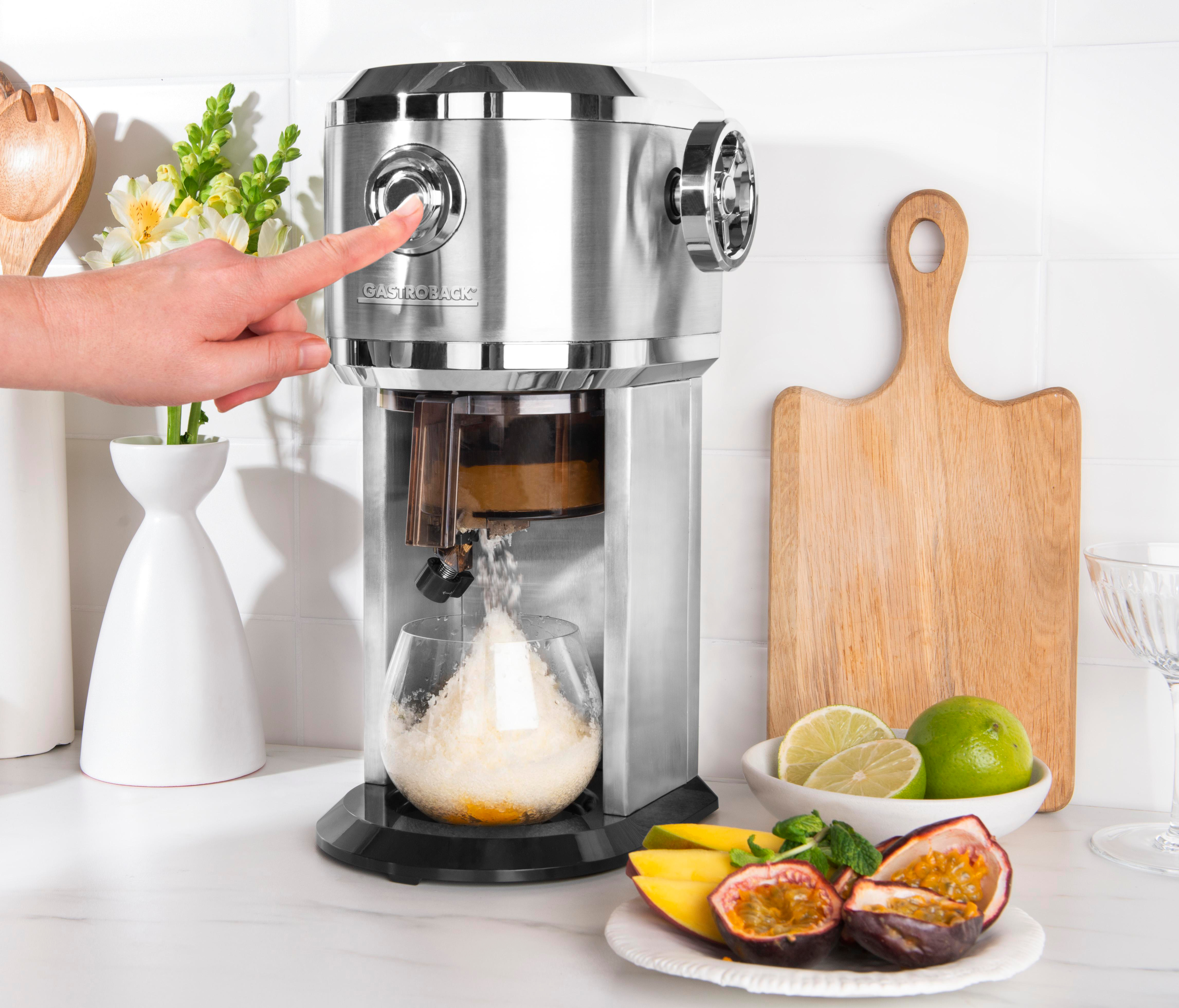 Eine Hand drückt einen Knopf an einem silbernen Entsafter. Ein Glas füllt sich mit Eis. Limetten, Mango und Passionsfrucht sind auf einem Teller.